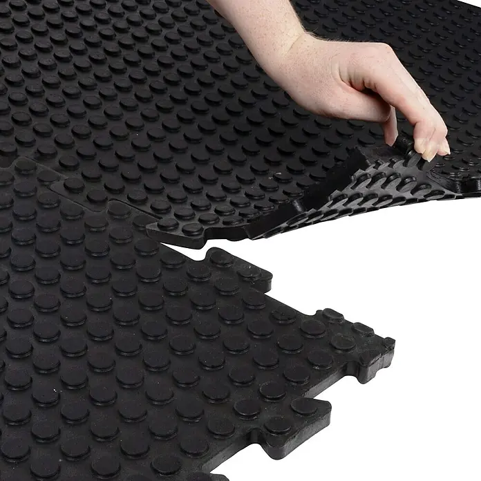 Floordirekt Bodenschutzmatte Puzzle Gummiläufer Buffallo - 100 x 100 cm, Schwarz - Ein schwarzer Gummibodenbelag mit Noppen wird von einer Hand angehoben, um das Stecksystem zu zeigen. Ideal für Fitnessstudios und Werkstätten.