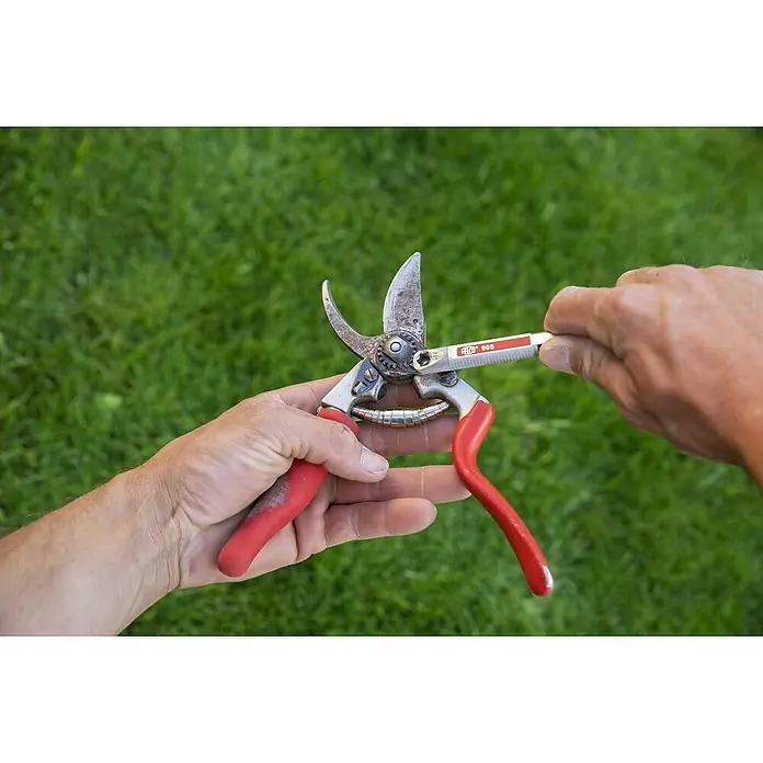 Felco Universal-Schleifgerät - Passend für: Felco Baum-, Reb- und Gartenscheren und Bypass-Astscheren für Rechtshänder - Bild einer Gartenschere mit roten Griffen, die mit einem FELCO 905 Schärfwerkzeug geschärft wird, vor grünem Gras.