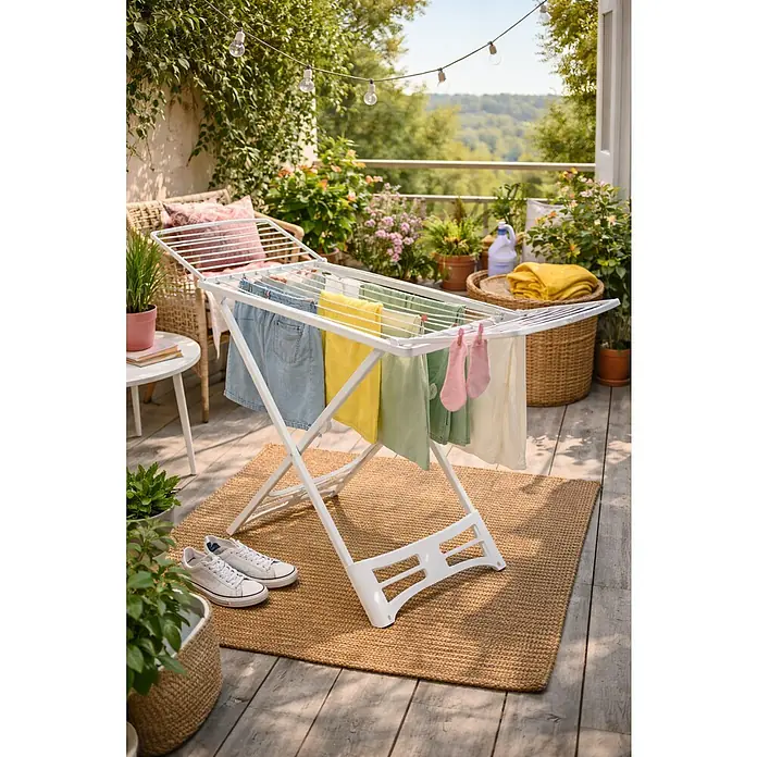 Tendedero con alas P-2/A Resina - Longitud cuerda de tender: 20 m, L x An x Al: 182 x 55 x 88 cm, Blanco - Tendedero en un balcón con ropa colgada para secar. El tendedero es blanco y está colocado sobre una alfombra. Se pueden ver plantas al fondo.