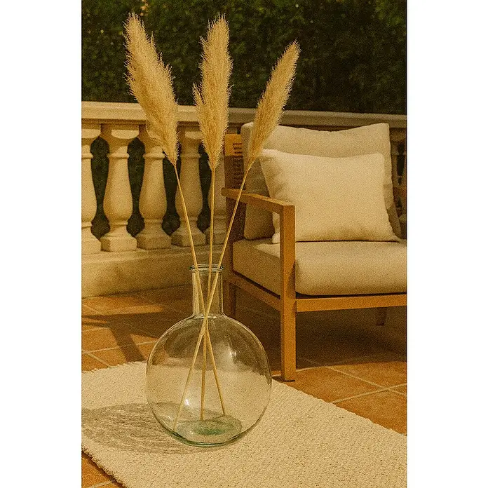 Jarrón Lilou - Ø x Al: 18 x 24 cm, Vidrio, Transparente - Primer plano de un jarrón de cristal con tres tallos altos de hierba de la pampa dorada sobre una alfombra beige frente a un sillón.