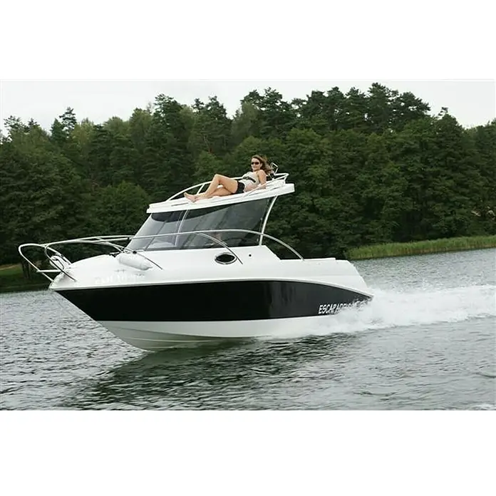 PEGAZUS GFK-Boot Escapade 600 Base SunTop - Max. 6 Personen, Nutzlast: 660 kg, CE-Klasse C - Eine luxuriöse Motoryacht mit einer Frau, die sich sonnt, fährt über einen ruhigen See, umgeben von üppigem Grün. Das Boot ist weiß mit schwarzen Akzenten.