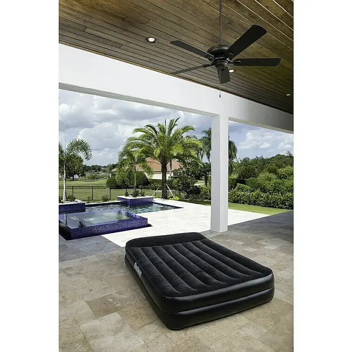 Bestway Luftbett Premium+ - 203 x 152 x 46 cm, Ausstattung: Integrierte Luftpumpe - Eine schwarze Bestway Luftmatratze liegt auf einem Steinboden im Freien, mit Pool und Palmen im Hintergrund. Ideal zum Entspannen!