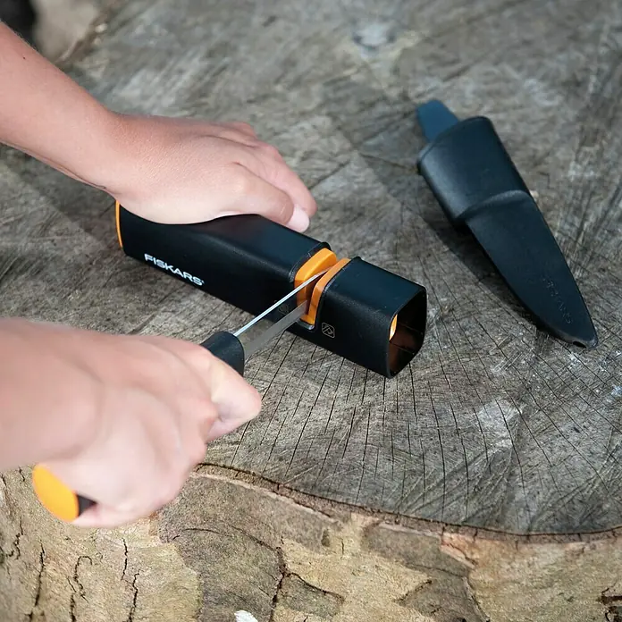Fiskars Axt- & Messerschärfer XSharp - Länge: 16,5 cm, Kunststoff - Nahaufnahme eines Fiskars-Messerschärfers auf einer Baumscheibe, während eine Person ein Messer schärft.