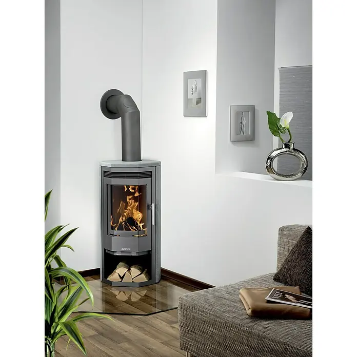 Justus Kaminofen Usedom 5 II - 5,5 kW, Raumheizvermögen: 88 m³, Material Verkleidung: Speckstein, Gussgrau/Speckstein - Ein moderner Justus Kaminofen mit Holzscheiten beheizt ein Wohnzimmer mit hellen Wänden, Kunstwerken und einem grauen Sofa.
