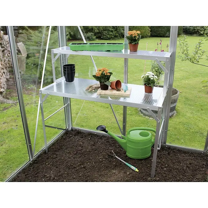 Vitavia Aufsatz für Gewächshaus-Tisch II - 28 x 121 x 39 cm, Aluminium - Ein Gewächshaus mit einem silbernen Anzuchttisch, auf dem Pflanzen, Töpfe und Gartengeräte stehen, darunter eine grüne Gießkanne auf der Erde.