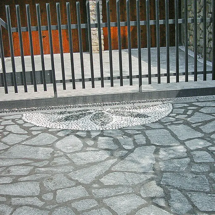 Polygonalplatte - Quarzit, Lieferumfang: ca. 10 m² - Detailaufnahme eines kunstvoll gestalteten Pflasters mit einem dekorativen Kieselmosaik vor einem modernen Metallzaun.