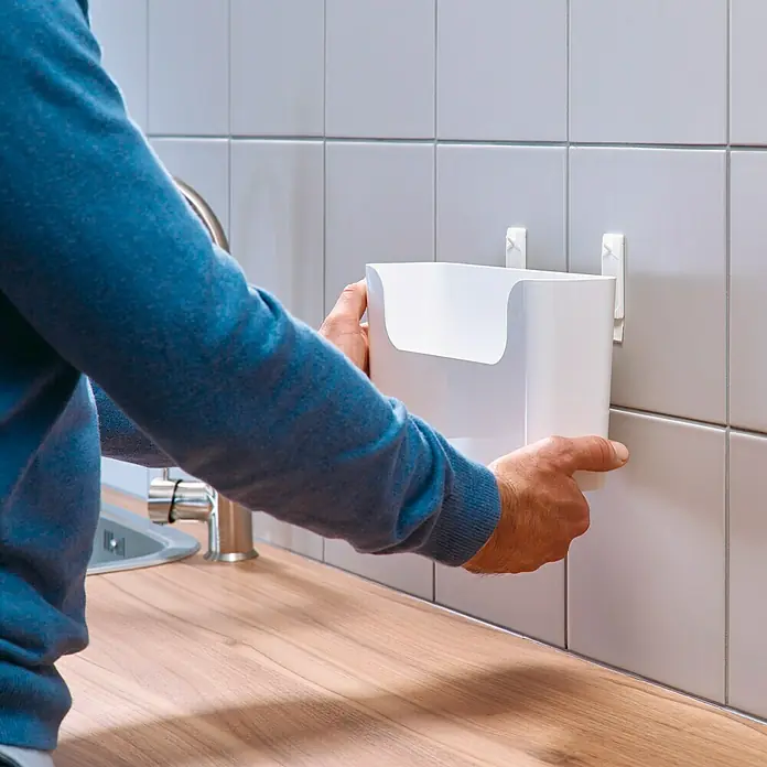Tesa Clavo adhesivo - Blanco, Carga soportada: 3 kg, 2 ud. - Un hombre está fijando un porta utensilios de cocina blanco a la pared. El accesorio de cocina proporciona espacio de almacenamiento y organización.