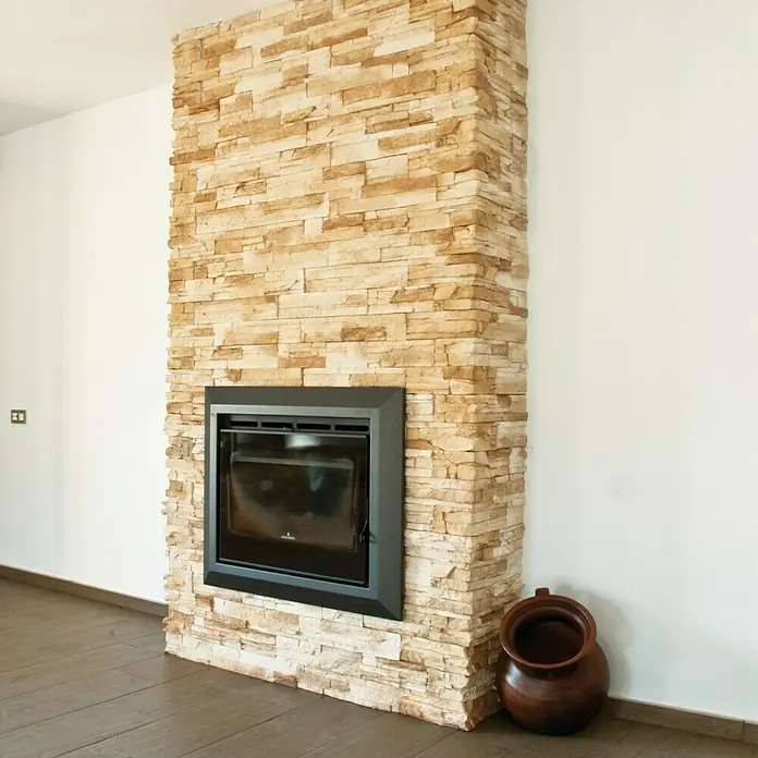 Revestimiento de piedra Petra Arena - Cantidad total: 4 pzs., Arena - Primer plano de una chimenea revestida de piedra con una jarra al lado, que añade un toque rústico a la habitación.