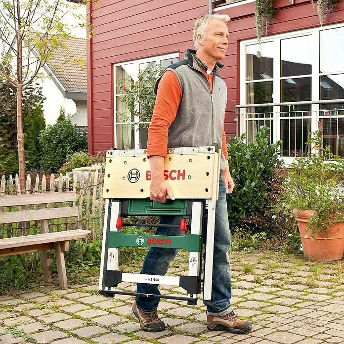 Bosch Werkbank PWB 600 - L x B: 680 x 680 mm, Arbeitshöhe: 834 mm, Belastbarkeit: 200 kg - Ein Mann trägt eine zusammengeklappte Bosch PWB 600 Werkbank im Garten. Die mobile Werkbank ist leicht und einfach zu transportieren.