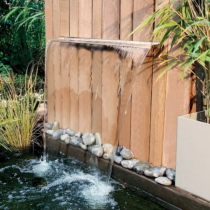 Ubbink Einbau-Wasserfall Niagara LED 90 Wall - 20 x 90 x 10 cm, Edelstahl, Leuchtmittel: LED - Nahaufnahme eines Wasserfall-Gartenbrunnens, der an einer Holzwand befestigt ist und in einen mit Steinen gesäumten Teich mündet.