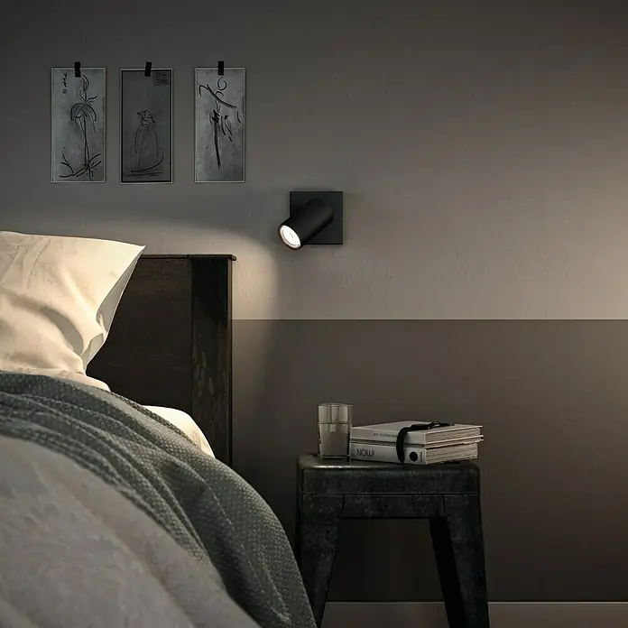 Philips Foco de pared Kosipo - 10 W, L x An x Al: 10,2 x 10,2 x 13,4 cm, Negro, GU10 - Foto moderna de un dormitorio con un foco negro de pared sobre una mesita de noche, emitiendo una luz cálida.