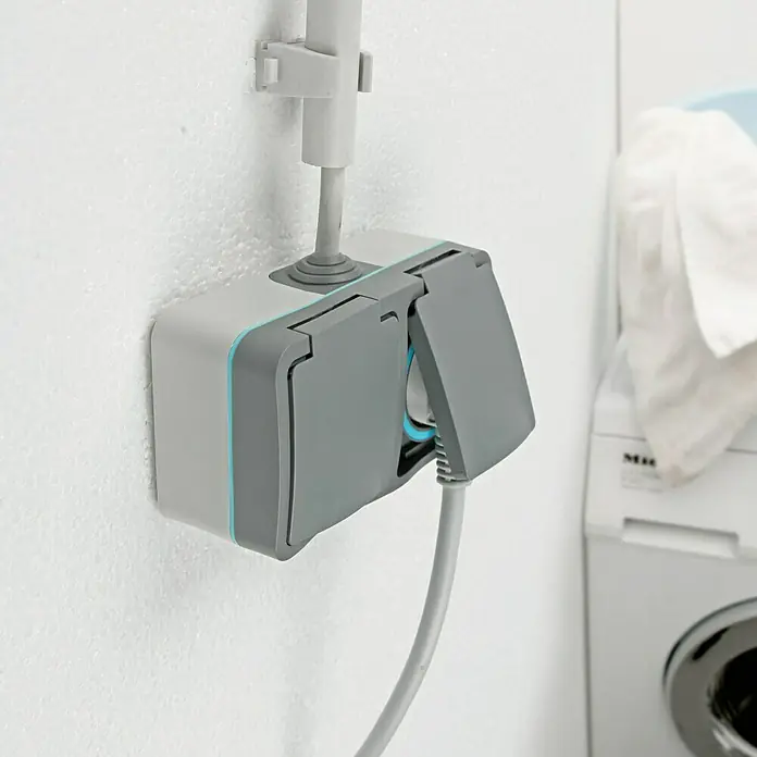 Voltomat Blueline Feuchtraum-Steckdose - 2-fach, Horizontal, IP54, Grau - Detailaufnahme einer spritzwassergeschützten Steckdose mit grauem Kabel und hellgrauem Gehäuse an einer weißen Wand.