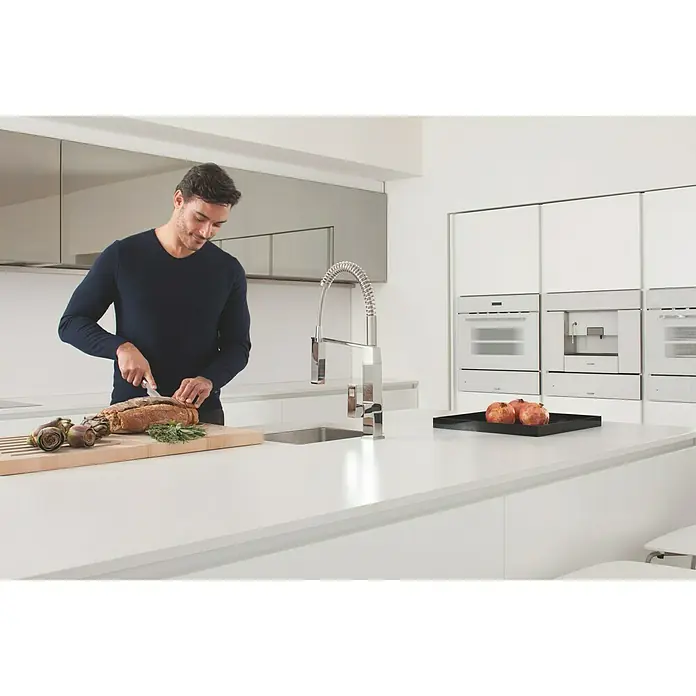 Grohe Eurocube Spültischarmatur - Chrom, Glänzend, Anzahl Strahlarten: 2 Stk. - Mann schneidet Brot an einer modernen Küchenspüle mit Chromarmatur, während im Hintergrund Einbaugeräte zu sehen sind.