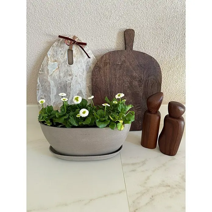 Ecopots Sofia Pflanzschale rund Oval - Grau - Stillleben mit Topfblumen, Schneidebrettern und Holzfiguren auf einer hellen Oberfläche, das die rustikale Ästhetik hervorhebt.