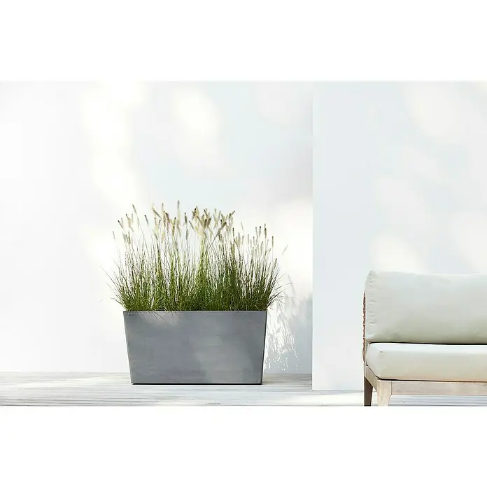 Ecopots Pflanzkasten Paris - Außenmaß (L x B x H): 100,5 x 39,6 x 51,8 cm, Kunststoff, Grau, Ausstattung Pflanzgefäß: Rollen - Pflanzkasten aus grauem Beton mit hohem Gras, neben einem hellen Outdoor-Sofa auf einer Terrasse mit weißer Wand.