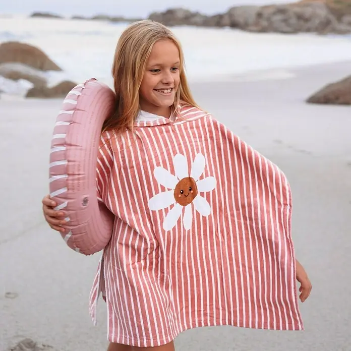 Swim Essentials Badeponcho - Baumwolle, Rot-Weiß - Mädchen am Strand mit einem gestreiften Poncho mit Gänseblümchen-Motiv, das ein hellrosafarbenes aufblasbares Schwimmbecken hält.
