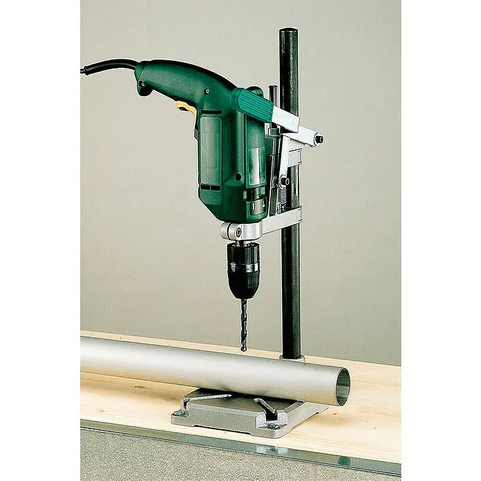 Wolfcraft Soporte de taladro - L x An x Al: 160 x 230 x 500 mm, Carrera: 60 mm, Apertura máxima: 112 mm - Un taladro verde está montado en un soporte y está haciendo un agujero en un tubo plateado que reposa sobre una superficie de madera.