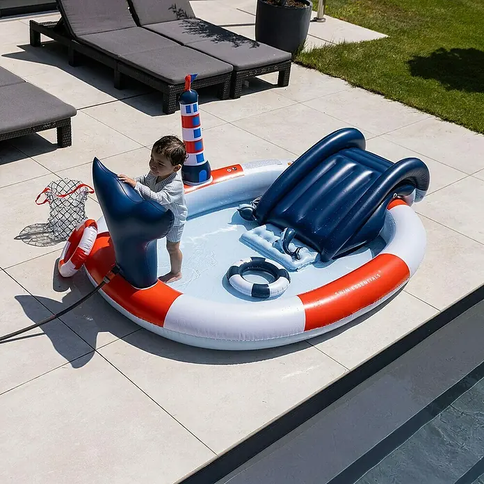 Swim Essentials Planschbecken Whale - Ø x H: 210 x 150 cm - Ein aufblasbares Planschbecken mit Rutsche und Leuchtturm steht auf einer Terrasse, ein Kind spielt darin.