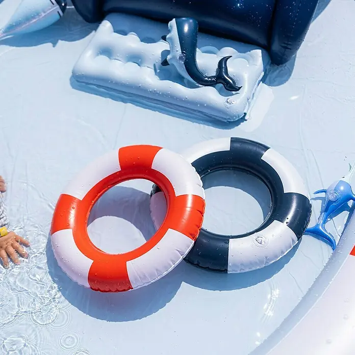 Swim Essentials Planschbecken Whale - Ø x H: 210 x 150 cm - Zwei Schwimmreifen (rot-weiß und schwarz-weiß) liegen im Wasser, umgeben von Wasserspielzeug und einem aufblasbaren Wal.
