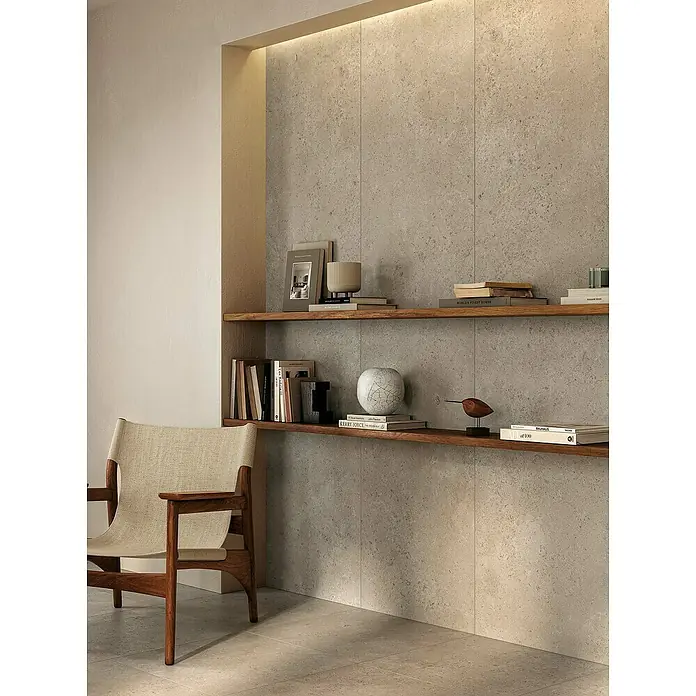 Atlas Concorde Solution Feinsteinzeugfliese Lunastone - 60 x 120 cm, Ash, Matt - Das Bild zeigt ein modernes Interieur mit einem hellen Sessel, einem Regal mit Büchern und Deko-Elementen vor einer strukturierten Wand.