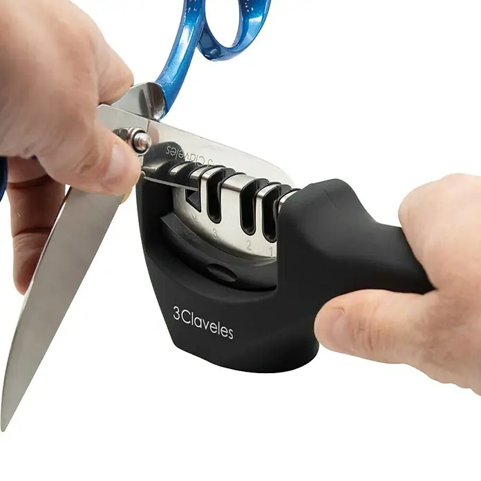 3 Claveles Afilador de cuchillos - Negro, Específico para: Cuchillos y tijeras - Un afilador de cuchillos 3Claveles siendo utilizado para afilar unas tijeras, con la hoja pasando por el aparato de afilar.