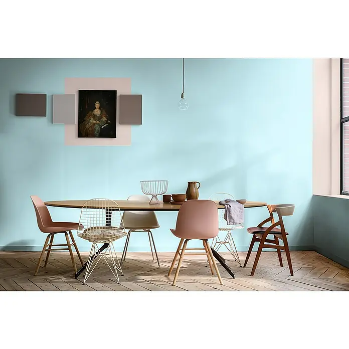Bruguer Elegance Pintura para paredes - Azul aqua, Mate, 4 l - Un comedor con una mesa ovalada y sillas de colores contrastados. Obras de arte decoran la pared, iluminadas por una sola bombilla.