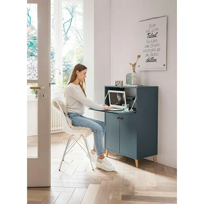 Schildmeyer Rena Sekretär - L x B x H: 39 x 65 x 102 cm, Fjord - Eine Frau sitzt an einem blauen Sekretär mit einem Laptop. Der Sekretär hat Holzfüße und einen ausklappbaren Schreibtisch.