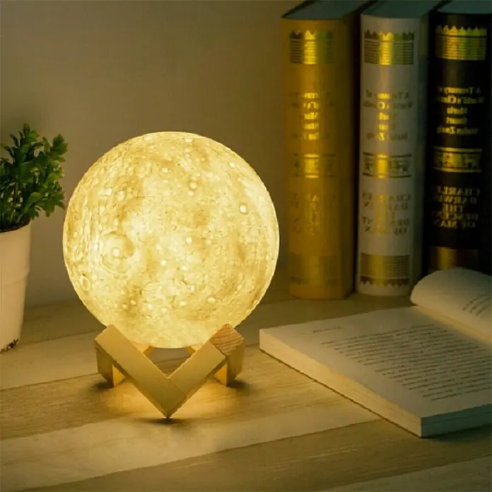 Voltomat Tischleuchte Luna - 15 W, Weiß, RGB - Ein leuchtender Mondlampe mit Holzsockel steht auf einem Tisch neben Büchern und einer Pflanze.