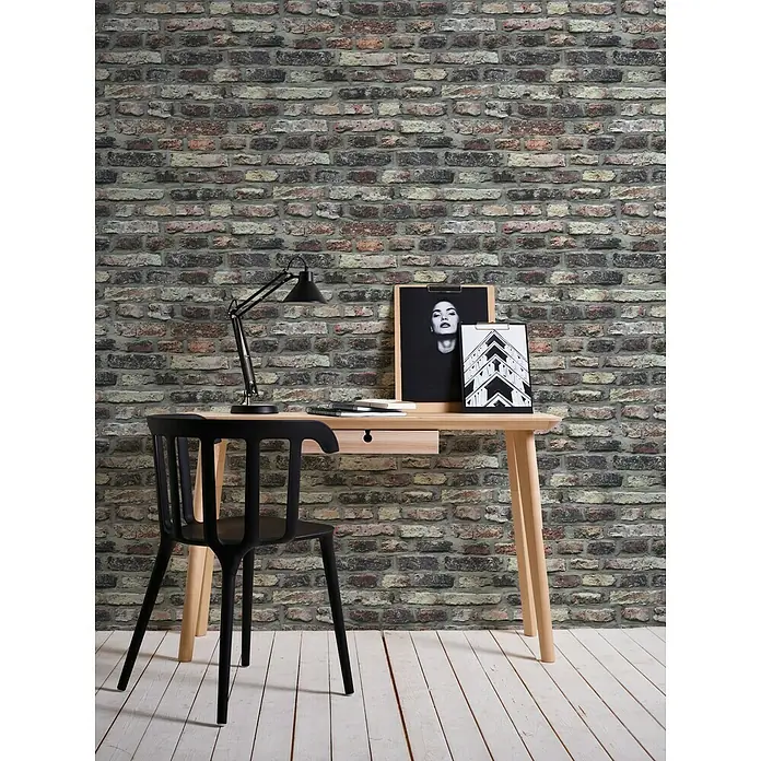 A.S. Création Il Decoro Vliestapete Steine - Grau/Rot, Steinoptik, 10,05 x 0,53 m - Ein Arbeitsbereich mit einem hellen Holzschreibtisch, einem schwarzen Stuhl, einer Lampe und gerahmten Bildern vor einer gemusterten Ziegelwand.