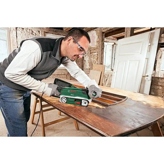 Bosch Elektro-Bandschleifmaschine PBS 75 A - 710 W, Bandgeschwindigkeit: 300 m/min - Mann schleift eine Tischplatte mit einem grünen Bosch Bandschleifer. Staubschutz und Schutzkleidung sind vorhanden.
