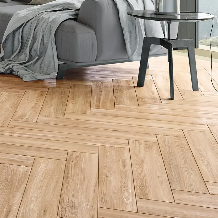 Steingutfliese Mini York - 60 x 11 cm, Honey, Matt - Nahaufnahme von hellbraunem Chevron-Parkettboden unter einem grauen Sofa und Beistelltisch, das warme und einladende Ambiente hervorhebt.