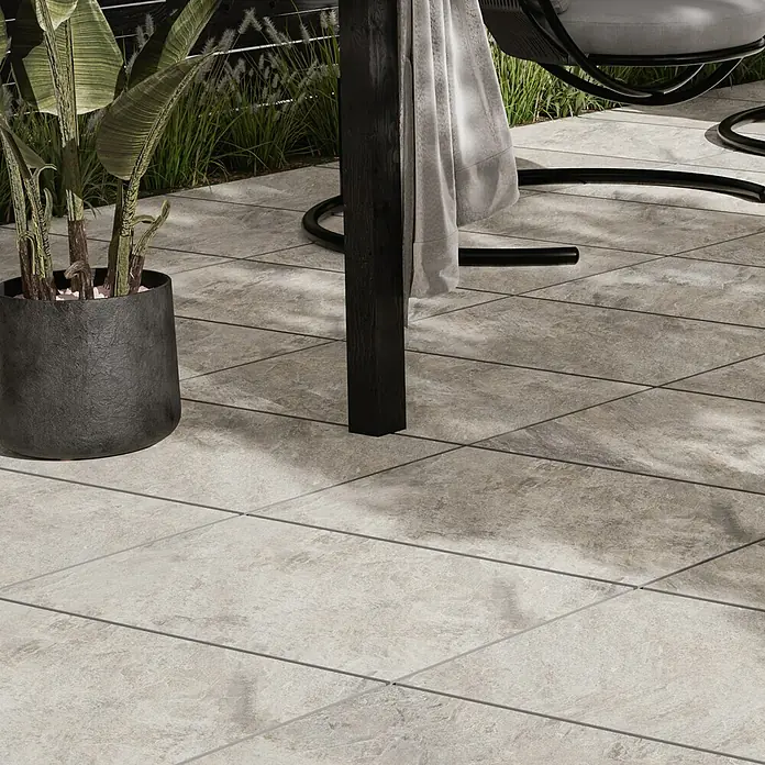 Terrassenfliese Homecraft - 59,7 x 119,7 x 2 cm, Grey, Matt - Nahaufnahme von beigen Terrassenfliesen mit einer Topfpflanze und einem Teil eines Stuhls im Hintergrund.