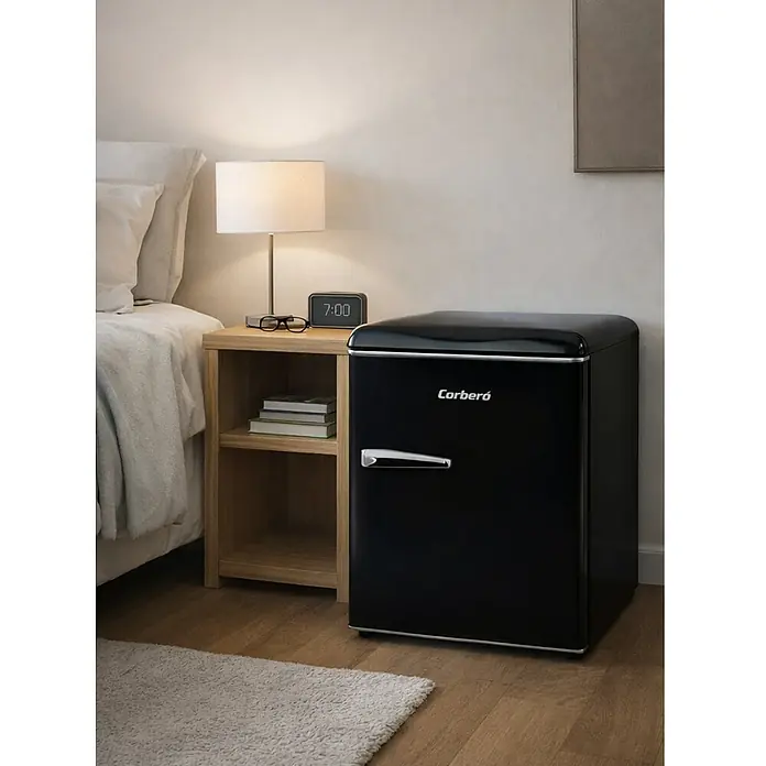 Corberó Frigorífico CFGMB45RN - Contenido útil total: 45 l, 53,4 cm x 44,7 cm x 55,5 mm, Negro - Primer plano de un mini frigorífico negro Corberó junto a una mesita de noche con lámpara, gafas y reloj en un dormitorio.