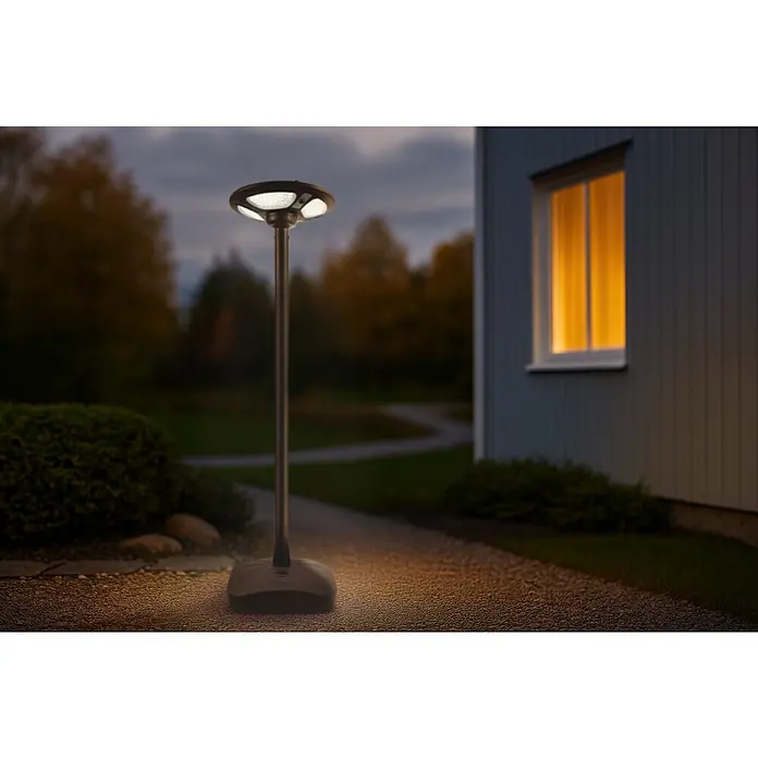 Garza Farola solar Icaro - Altura: 265,5 cm, Negro, IP54, Blanco cálido - Una lámpara exterior moderna en un camino de grava, que ilumina el paseo hacia la casa. Al fondo, una ventana brilla con luz cálida.