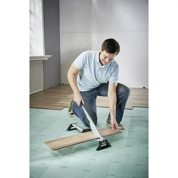 Wolfcraft Cortaláminas de parquet LC 600 - Ancho de corte: 465 mm, Aluminio - Un hombre se arrodilla mientras corta una tabla de laminado con un cortador Wolfcraft, asegurando un ajuste perfecto para el pavimento.
