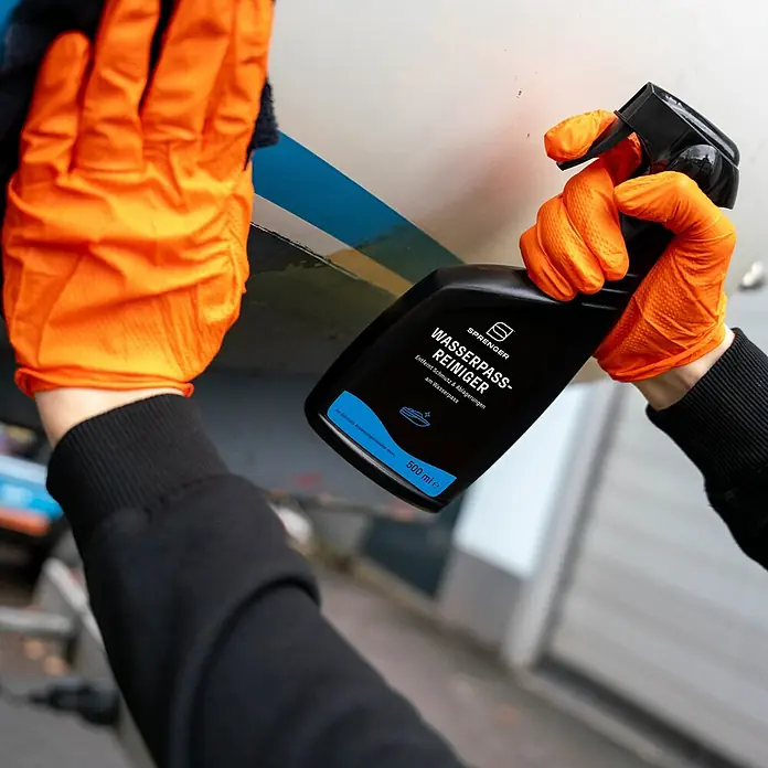 Sprenger Wasserpassreiniger - 500 ml - Das Bild zeigt eine Person, die einen Sprenger Wasserpass-Reiniger mit orangefarbenen Handschuhen verwendet, um ein Boot zu reinigen.