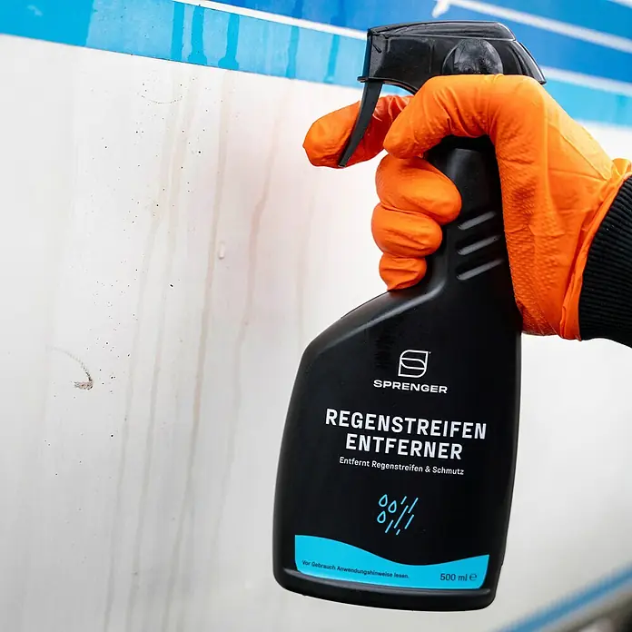 Sprenger Regenstreifen-Entferner - 500 ml - Eine Hand in einem orangefarbenen Handschuh hält eine schwarze Sprühflasche "Regenstreifen Entferner" vor einer verschmutzten Oberfläche.