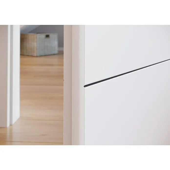Kilsgaard Zimmertür 14/14 - 735 x 1.985 mm, DIN Anschlag: Links, Weiß lackiert, ähnlich RAL 9010, Röhrenspan, Schwarze V-Fugen - Nahaufnahme einer minimalistischen weißen Tür mit einem schlanken schwarzen Griff und einem Blick auf einen hellen, holzgetäfelten Flur.