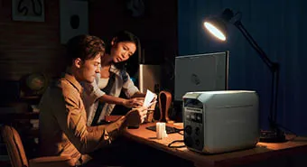 Frau und Mann sitzen im Büro am Laptop mit einer Powerstation und einer eingeschalteten Tischlampe