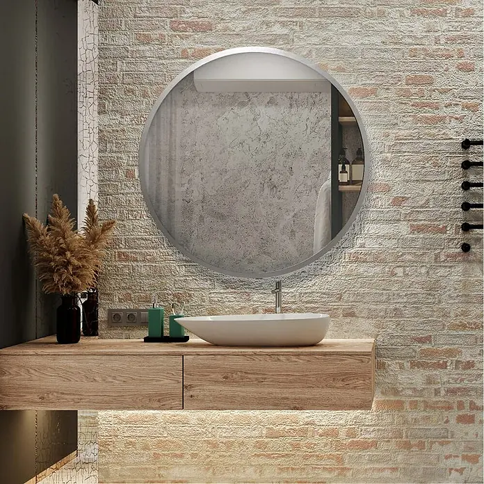 Espejo de pared Circular - Diámetro: 55 cm, Plata betún - Un espejo redondo cuelga sobre un tocador con cajones de madera, iluminado desde abajo, contra una pared de ladrillos. Diseño de baño elegante.