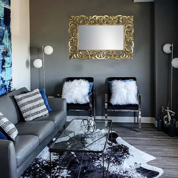 Espejo de pared Berrocal - 88 x 128 cm, Oro betún - Elegante sala de estar con un espejo dorado sobre dos sillones, acentuada por un sofá moderno y una mesa de centro elegante.