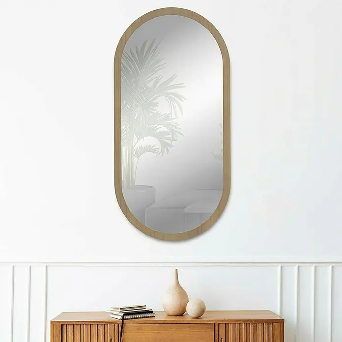 Espejo de pared Alba - 58 x 118 cm, Roble - Un espejo decorativo ovalado con marco de madera clara cuelga sobre un mueble de madera, con jarrones y libros encima.