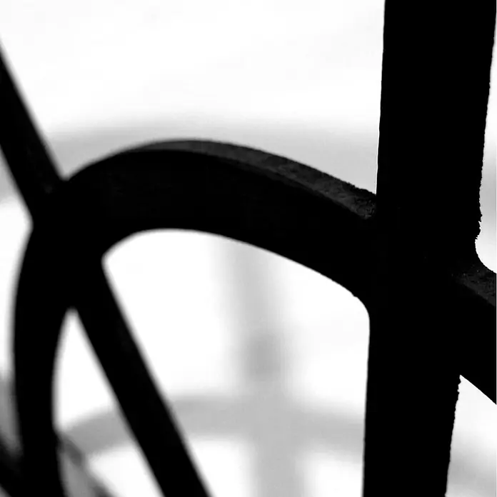 Espejo de pared Catania - 70 x 100 cm, Negro - Primer plano de una estructura metálica negra ornamentada sobre un fondo brillante y borroso.