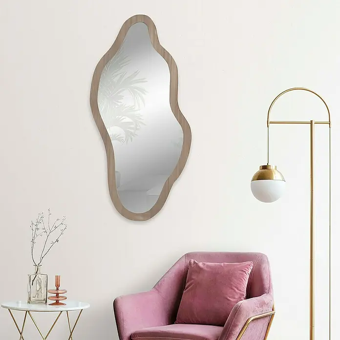 Espejo de pared Onda - 38 x 78 cm, Roble - Un espejo de forma irregular con marco de madera cuelga de una pared, complementado con un sillón y una lámpara de pie.
