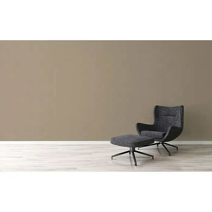 A.S. Création Ethnic Origin Vliestapete - Braun, Uni, 10,05 x 0,53 m - Grauer Sessel mit Hocker vor einer hellbraunen Wand auf einem hellen Holzboden. Bequeme und stilvolle Sitzgelegenheit für entspannte Momente.