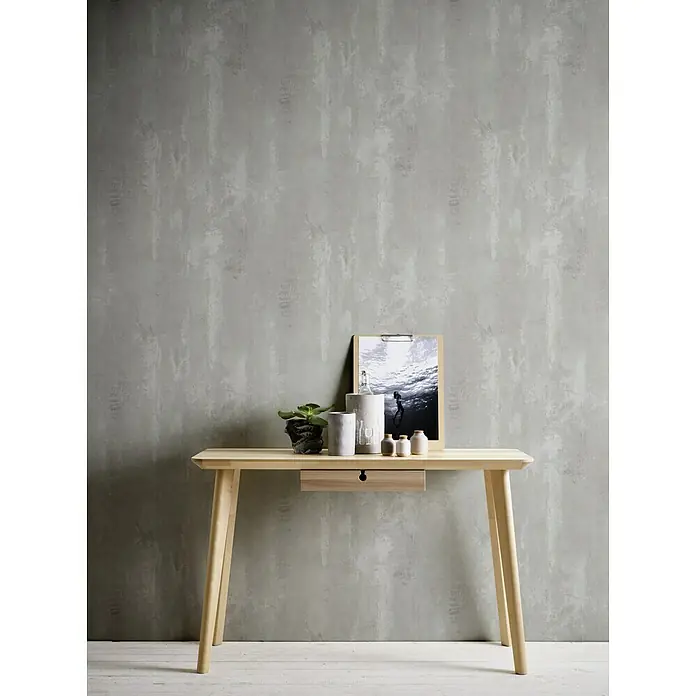 A.S. Création Neue Bude 2.0 ED II Vliestapete Concret - Grau, Betonoptik, 10,05 x 0,53 m - Eine helle Holzkonsole mit Schublade, dekoriert mit Pflanzen, Keramik und einem gerahmten Bild, vor einer grauen Wand.