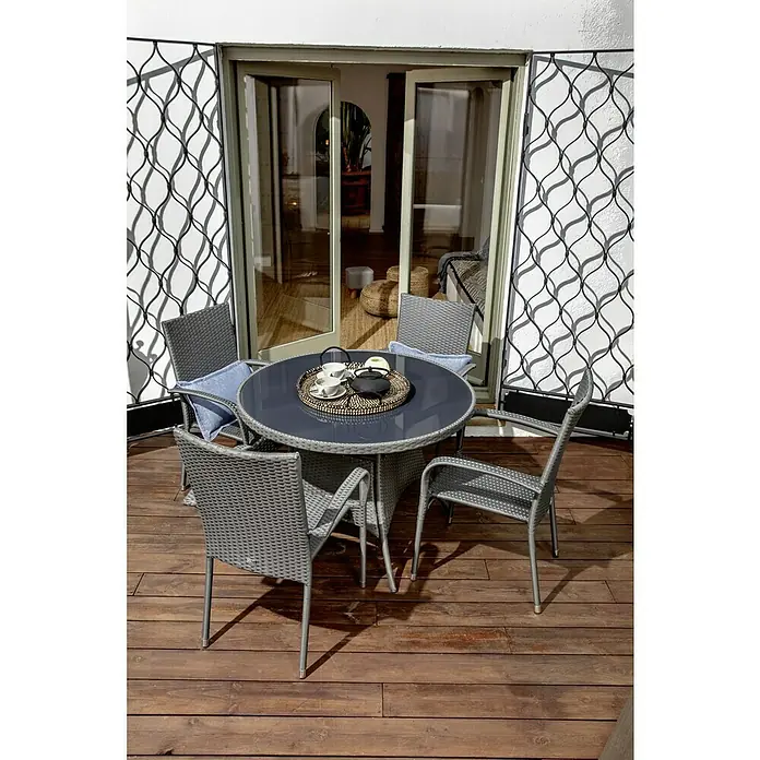 Sunfun Neila Sillón de jardín Neila - An x Pr x Al: 56 x 63 x 94 cm, Poliratán, Apilable, Gris plata - Un conjunto de patio gris con una mesa redonda y cuatro sillas se sitúa en una terraza de madera delante de una puerta de patio abierta.