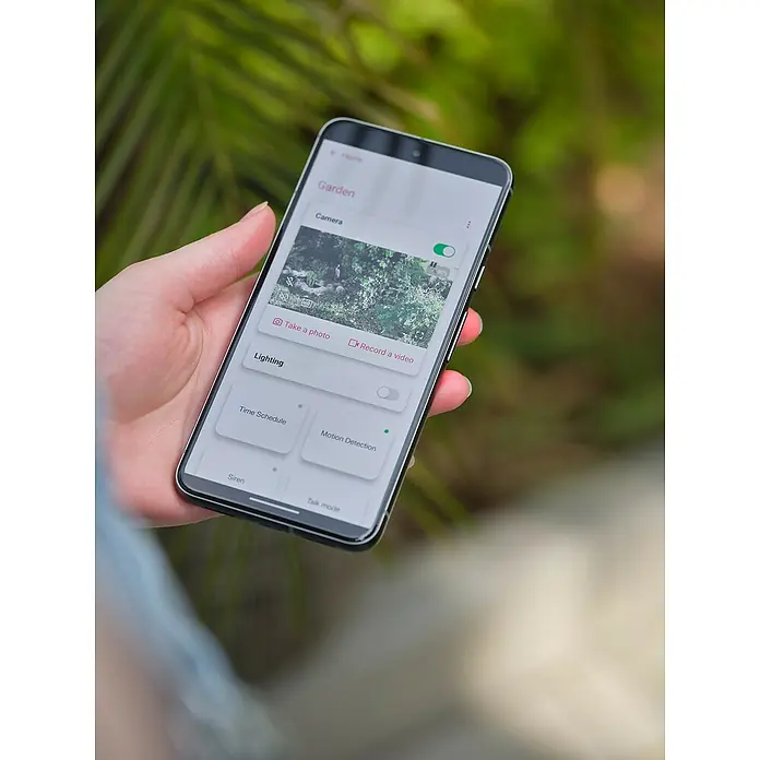 Osram Smart+ Überwachungskamera Battery Solar - Weiß - Eine Hand hält ein Smartphone, das eine Sicherheitskamera-App mit Gartenansicht und verschiedenen Steuerungsoptionen anzeigt.