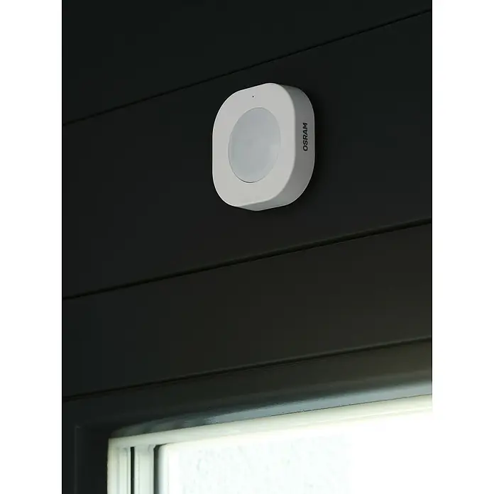 Osram Smart+ Bewegungsmelder - Weiß - Ein weißer OSRAM-Bewegungsmelder ist an einer dunklen Wand montiert und sorgt für automatisches Licht bei Bewegungserkennung.