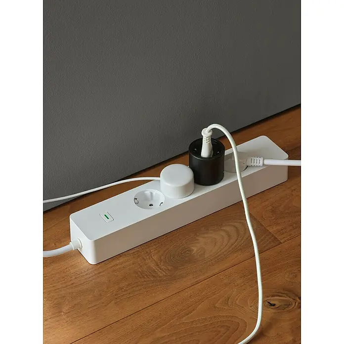 Osram Smart+ Smart-Steckdose matter - Innen, Schwarz, 1 Stk. - Eine weiße Steckdosenleiste mit mehreren Anschlüssen, zwei davon belegt, liegt auf einem Holzboden vor einer grauen Wand.